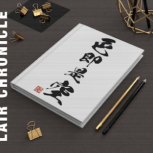 May include: White journal with black Japanese calligraphy and a red stamp. Gold and black pencils and gold binder clips are arranged around the journal. The text "LAIR CHRONICLE" is on the left side.