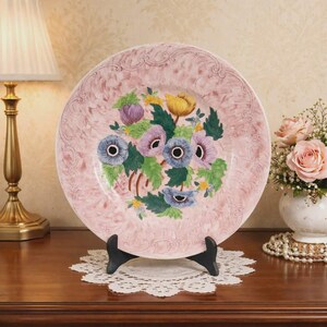 May include: A pink decorative plate with a floral design. The plate features a bouquet of colorful flowers in the centre, including purple, blue, yellow, and pink blooms. The plate is displayed on a black stand, with a lamp and vase of flowers in the background.