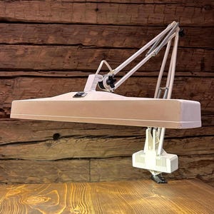 Puede incluir: Una lámpara de escritorio blanca y ajustable con una luminaria rectangular. La lámpara tiene una serie de brazos articulados y una base de abrazadera. La lámpara es de metal y plástico.