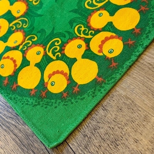 Puede incluir: Una tela verde con un patrón repetido de pollitos amarillos con patas y crestas rojas. Los pollitos están dispuestos en un patrón circular con remolinos decorativos. La tela está sobre una superficie de madera.