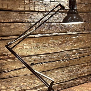 Puede incluir: Una lámpara de escritorio negra ajustable con una pantalla en forma de cúpula. La lámpara tiene un brazo largo y articulado con un mecanismo de resorte y una base rectangular. La lámpara está colocada contra una pared de madera.