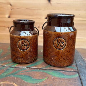 Puede incluir: Dos botes de cerámica marrón con tapas y asas de color marrón oscuro. El bote más grande tiene una marca de "1/3", y el más pequeño tiene una marca de "1/4". Ambos tienen un acabado texturizado y moteado, probablemente para uso en la cocina.