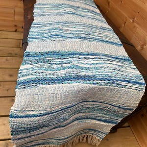 Puede incluir: Una alfombra corredora tejida con un patrón de rayas azules y blancas. La alfombra tiene un borde con flecos y está hecha de un material texturizado. La paleta de colores incluye tonos de azul, blanco y gris.