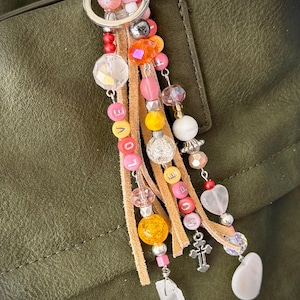 May include: A colorful key chain with a variety of beads, charms, and tassels. The key chain features a silver ring, pink, orange, yellow, and white beads, a silver cross charm, a pink heart charm, and a white stone charm. The key chain also has a leather tassel with the word "LOVE" spelled out in pink and yellow beads.