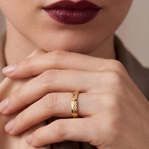 May include: A gold band ring featuring a repeating chevron or herringbone pattern engraved along the entire outer surface. The ring has a polished metallic finish and a consistent width, showcasing a detailed texture that wraps around the finger.