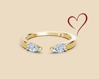 May include: A gold ring with two pear-shaped diamonds set on either side of the open band. The ring is on a beige background with a red heart drawn in the upper right corner. The ring is a piece of jewelry.