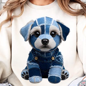 May include: A plush puppy toy made of denim fabric, wearing denim overalls. The puppy has large, expressive eyes and a stitched design. The toy is sitting on a white background. The Paper and Play logo is in the bottom right corner.