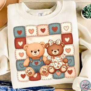 May include: Cream-colored sweatshirt featuring a colorful graphic of two teddy bears. One bear wears denim overalls, the other a floral dress. The background is a patchwork of squares with heart designs.