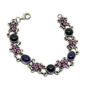 May include: Silver-toned bracelet featuring alternating dark purple and oval-shaped purple gemstones, interspersed with clusters of small pink stones. The bracelet has a lobster clasp closure and is set against a white background.