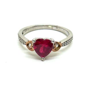May include: A silver ring with a heart-shaped, deep red gemstone. The ring features gold-colored accents on either side of the heart and small, clear stones set along the band. The ring is a piece of jewelry.
