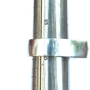 May include: A silver ring is placed on a ring sizer. The ring is a simple band design. The ring sizer is a metal cylinder with the number 5 visible. The ring is being measured for size.
