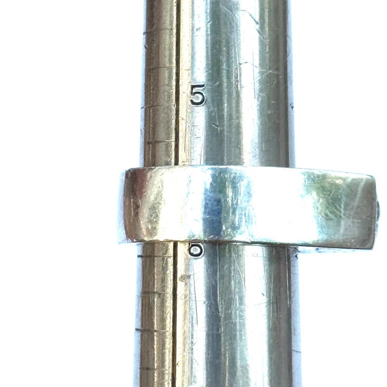 May include: A silver ring is placed on a ring sizer. The ring is a simple band design. The ring sizer is a metal cylinder with the number 5 visible. The ring is being measured for size.