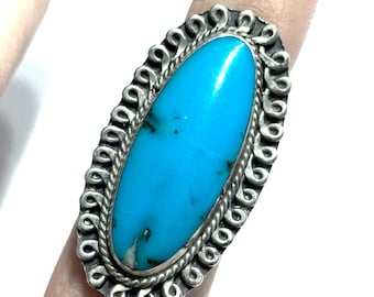 May include: Oval turquoise ring with a silver filigree border. The bright blue stone is the focal point, set in a detailed, ornate frame. The ring is shown on a finger, highlighting the size and design of the jewelry.