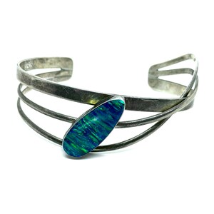 May include: A silver cuff bracelet with a vibrant, oval-shaped blue and green opal stone. The bracelet features a multi-strand design, with three parallel bands curving around the wrist. The opal stone is the focal point, set in a silver bezel.