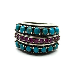 May include: A multi-band ring featuring three distinct rows of stones set in silver. The top and bottom rows consist of oval-shaped turquoise cabochons, while the center row contains a series of round, deep pinkish-red faceted gemstones.