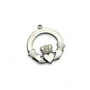 May include: Silver-toned Claddagh charm featuring a heart, crown, and hands. The heart is centered, topped with a crown, and clasped by two hands within a circular frame. The charm has a small loop at the top for attachment.