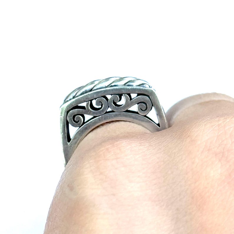 May include: A silver ring with a decorative design. The ring features a braided top and a detailed openwork pattern with swirling shapes. The ring is worn on a finger, showcasing its intricate craftsmanship and design.