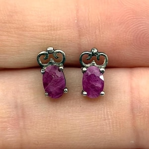 May include: A pair of stud earrings featuring oval-shaped, deep pinkish-red gemstones set in silver-toned metal. Each earring has a decorative, openwork scroll design at the top, positioned directly above the faceted, oval-cut stone setting.