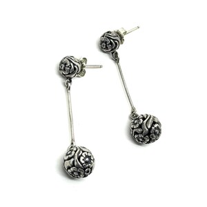 May include: A pair of silver dangle earrings. Each earring features a floral design on the stud and a dangling ball with a detailed floral pattern. The earrings are displayed against a white background.