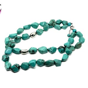 May include: A turquoise gemstone necklace with irregular, polished stones. The necklace features silver-toned metal accents and a clasp. The stones are a vibrant teal color with dark matrix patterns, strung on a black cord.