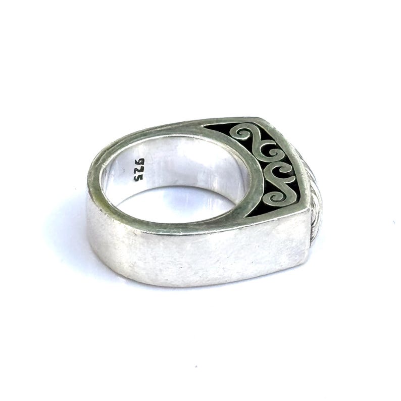 May include: A silver ring with a rectangular face and a decorative black and silver design. The ring is marked with "925" on the inside band. The ring has a brushed metal finish.