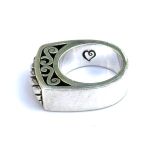 May include: A silver ring with a rectangular face featuring a black and silver scroll design. The ring has a heart symbol engraved on the inside. The ring is a size 8.