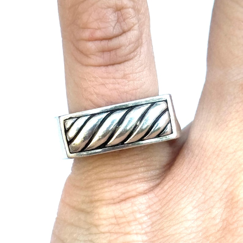May include: A silver-toned ring with a rectangular face featuring a twisted rope design. The ring is worn on a finger, showcasing its detailed craftsmanship and metallic sheen. The ring's design is a classic and versatile piece of jewelry.