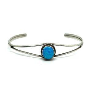 May include: A silver cuff bracelet with an oval, bright blue opal stone at the center. The bracelet has a delicate, open design with a thin band and a textured border around the stone. The opal has a vibrant, iridescent quality.