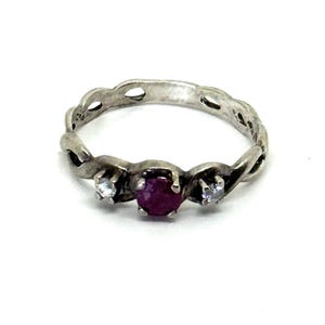 May include: A silver ring with a braided design, featuring a central round, deep red gemstone flanked by two smaller, clear gemstones. The ring is set against a white background, highlighting its intricate details.