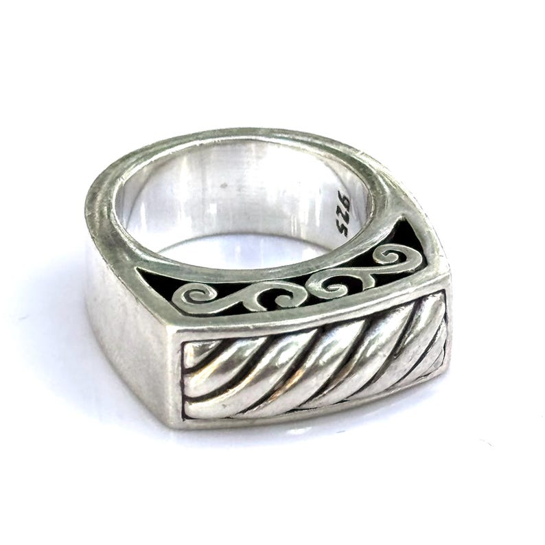 May include: A silver ring with a rectangular face. The ring features a twisted rope design on the bottom and a black inlay with a scroll design. The inside of the ring is stamped with "925".