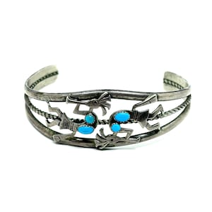 May include: Silver cuff bracelet featuring Kokopelli figures and turquoise stones. The bracelet has a three-strand design with twisted rope detailing and stylized figures. The turquoise stones are oval-shaped and add a pop of color to the silver.