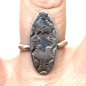 May include: Silver ring with an oval, engraved face. The ring features a detailed, symmetrical design with a central shape and surrounding patterns. The ring is worn on a finger, showcasing the intricate metalwork and vintage style.