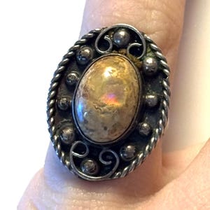 May include: Oval-shaped silver ring with a large, iridescent, beige stone. The ring features a detailed design with a twisted rope border, small silver balls, and decorative swirls. The stone has a polished, reflective surface.