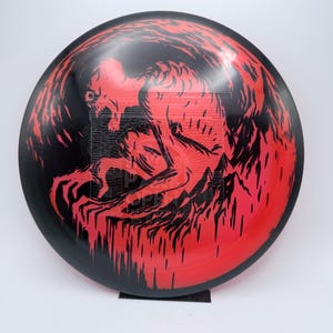 May include: A black and red disc golf disc with a graphic of a skeletal creature. The design shows a red figure with a menacing expression against a red background. The disc is circular and made of plastic.