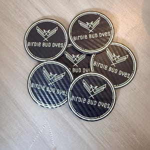 Könnte beinhalten: Eine Sammlung runder, schwarz-weißer Aufkleber. Jeder Aufkleber hat eine Kohlefaserstruktur und einen weißen Rand. Der Text "Birdie Bud Dyes" ist in Weiß gedruckt, zusammen mit einer stilisierten Vogelgrafik.