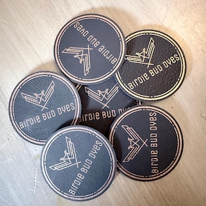 May include: A collection of black, circular stickers with a metallic rose gold border and logo. The logo features a stylized bird design and the text "Birdie Bud Dyes". The stickers are arranged on a light brown surface.