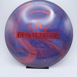 May include: A Dynamic Discs Raider disc golf disc with a marbled purple, blue, and red design. The disc features the word "RAIDER" in red, along with speed, glide, turn, and fade ratings. The text "DYNAMIC DISCS" is also visible.