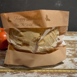 May include: A brown paper bag with a clear window, containing a loaf of bread. The bag is printed with "Happy Holidays" and "From: The Walsh Family" in a decorative font, along with holly leaf illustrations. The bag sits on a wooden surface.