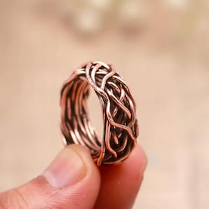 May include: A copper-colored ring featuring a complex, woven wire design that resembles a nest or tangled branches. The metal has a warm, metallic finish with darker, recessed areas that highlight the intricate, overlapping wire pattern.