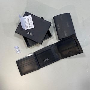 May include: A black leather BOSS wallet with multiple card slots and a coin pocket, displayed alongside its matching black presentation box. The wallet features the BOSS logo embossed on the interior and exterior, accompanied by a small silica gel packet.