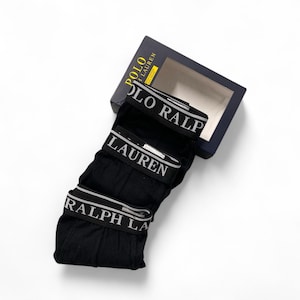 May include: A set of three black boxer briefs with a wide elastic waistband featuring the white text POLO RALPH LAUREN. The underwear is presented with a dark blue rectangular cardboard box that displays the brand name in yellow and white text.