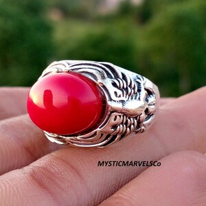 May include: A silver ring featuring a large, smooth, oval-shaped red stone set in a band with intricate, textured metal detailing. The text MYSTICMARVELSCO is visible on the lower right side of the ring, showcasing a bold and detailed design.
