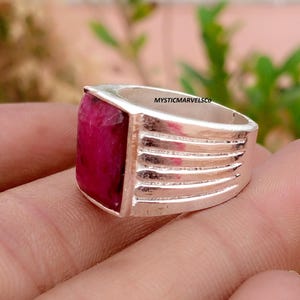 May include: A silver ring featuring a rectangular, deep pink ruby gemstone set in a polished metal band. The band displays a series of horizontal grooves along the sides. The text MYSTICMARVELSCO is visible on the inner surface of the ring.