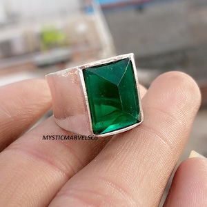 May include: A silver-toned ring featuring a large, rectangular, faceted emerald green gemstone set in a smooth metal band. The text MYSTICMARVELSCO is printed in black on the side of the silver band, which has a polished finish and a wide profile.