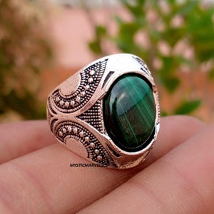 Könnte beinhalten: Ein silberfarbener Ring mit einem ovalen, dunkelgrünen Malachitstein mit natürlicher Bänderung. Die Metallschiene zeigt ein kompliziertes, geprägtes Muster aus kleinen Punkten und geschwungenen Linien. Der Text MYSTICMARVELSCO ist auf dem Ring zu sehen.