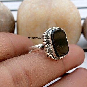 May include: A silver ring featuring a rectangular black stone set in a decorative frame with beaded and twisted metal detailing. The band is smooth and polished. The text MYSTICMARVELSCO is visible on the side of the ring setting.