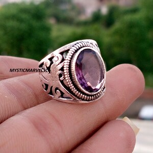 May include: A silver-toned ring featuring an oval-shaped purple gemstone set in a rope-textured bezel. The band has an openwork design with swirling patterns and the text MYSTICMARVELSO printed on the side of the metal surface.