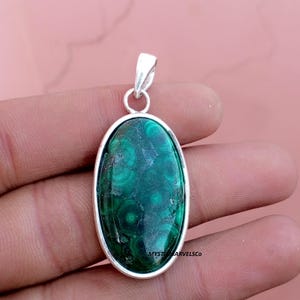 May include: Oval-shaped malachite pendant with a silver-colored frame. The malachite stone features a swirling pattern of dark and light green hues. The pendant has a small loop at the top for a chain.