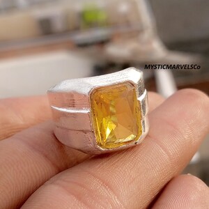 Op de afbeelding: Een zilverkleurige ring met een grote, rechthoekige, gefacetteerde gele edelsteen in het midden. De band heeft een brede, getextureerde, matte afwerking met drie horizontale ribbels aan elke kant. De tekst MYSTICMARVELSCo is zichtbaar in de hoek.