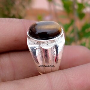 May include: A silver ring featuring a polished, oval-shaped brown tiger's eye gemstone set in a textured metal band. The band has a vertical grooved design and the text MYSTICMARVELSCO is visible on the side of the silver setting.